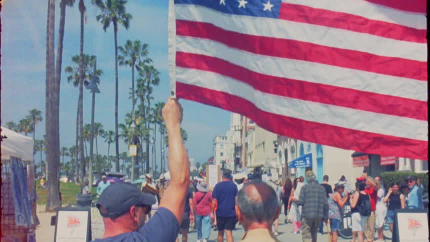 In a vintage-style clip, a man proudly carries a large American flag, leading a crowd along a sunny Venice Beach boardwalk