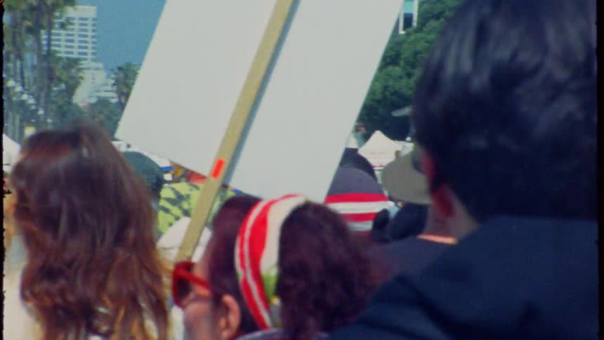 Vintage-style, behind-the-crowd shot of a political protest march with signs and an American flag waving