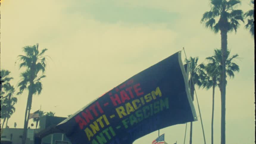 Vintage-style slow-motion footage of anti-hate and anti-Trump protest flags waving in the wind against a backdrop of palm trees