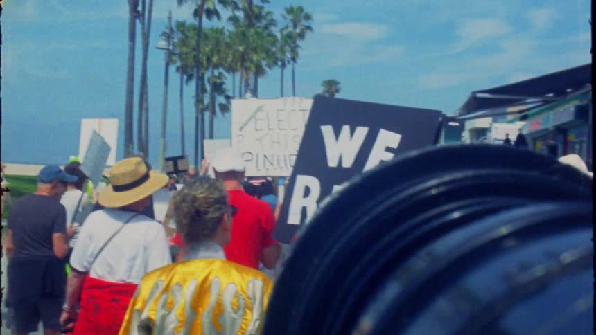 A crowd of activists march down a sunny, palm tree-lined street with signs, one prominently reading 'WE RISE,' in a vintage film style
