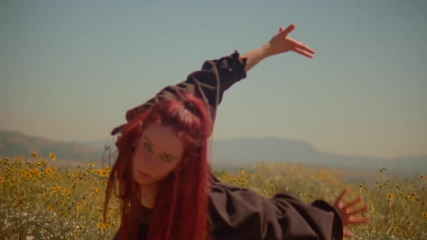 A young woman with vibrant red hair performs an expressive, slow-motion dance in a sunny field of yellow wildflowers