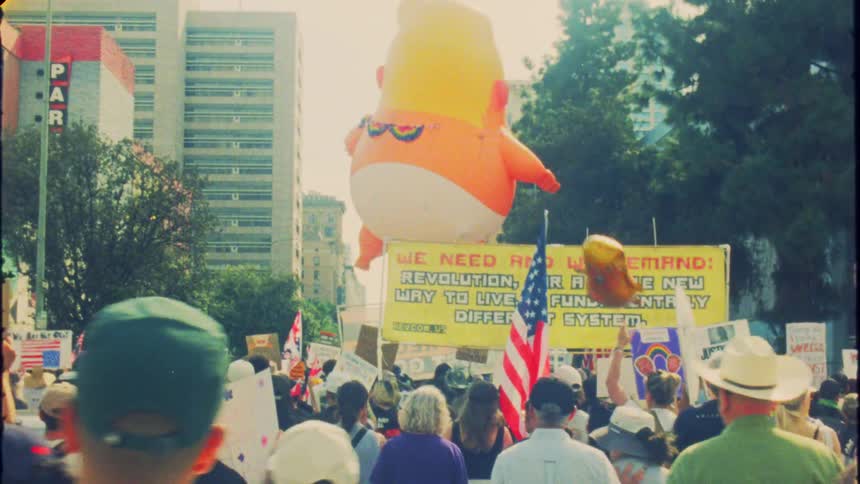 Vintage-style footage captures a large crowd marching in an anti-war protest featuring an inflatable 'Baby Trump' balloon and various signs