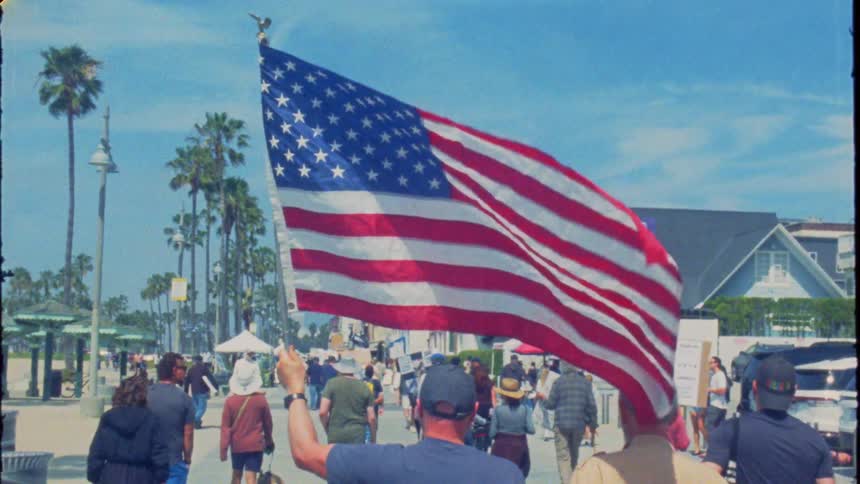 A slow-motion, vintage-style shot shows a man waving a large American flag while walking with a crowd on a sunny beach boardwalk
