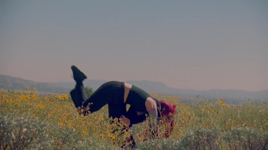 A woman with vibrant red hair performs an expressive and athletic dance in slow motion within a field of yellow wildflowers