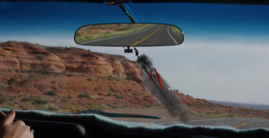 A decorative feather sways back and forth from a rearview mirror while driving on a scenic desert road