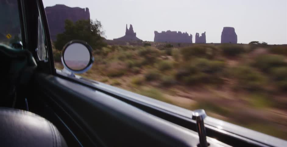 A classic car drives through the desert with Monument Valley's iconic buttes visible in the distance