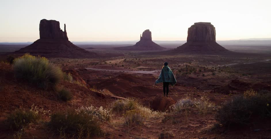 A person wrapped in a poncho stands on a dusty hill, looking out at the majestic buttes of Monument Valley at sunrise