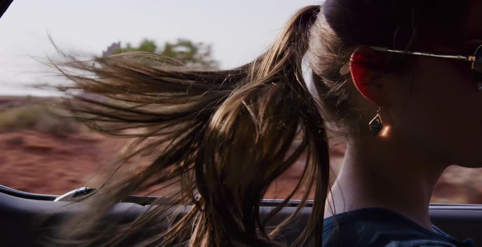 In slow motion, a woman's ponytail blows in the wind as she rides in a convertible on a road trip through Monument Valley