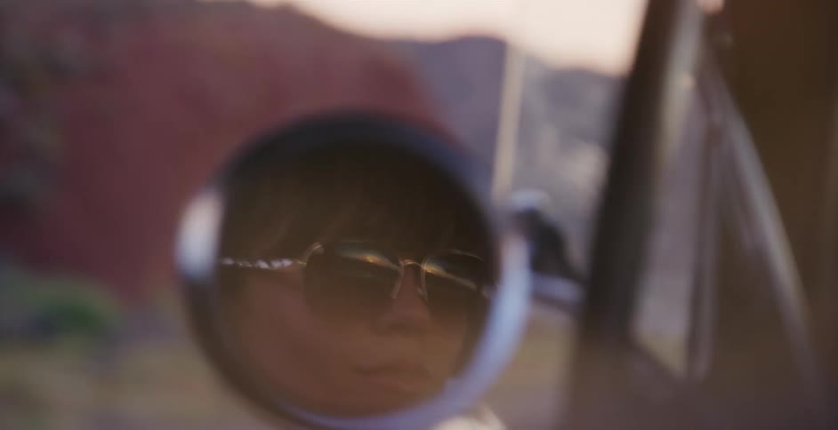 A woman's reflection is seen in the shaky side-view mirror of a car driving through a landscape at sunset