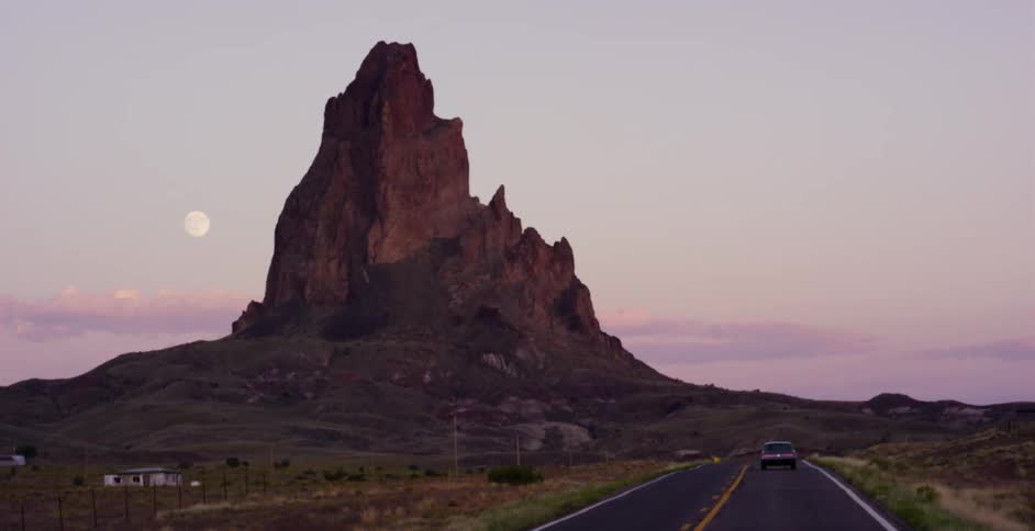 Cars drive on a remote desert road towards an iconic rock formation as the full moon rises during a beautiful sunset
