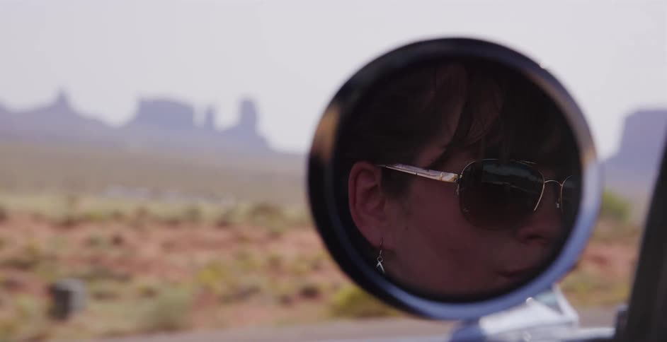 Close-up of a woman's reflection in a car's side-view mirror as she drives through the desert landscape of Monument Valley