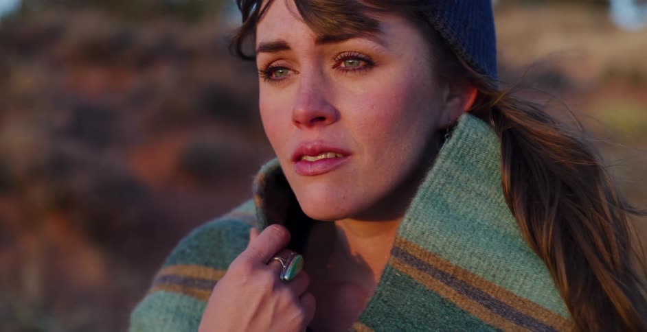 A close-up shot of a beautiful, emotional young woman wrapped in a warm blanket looking off into the distance at sunset