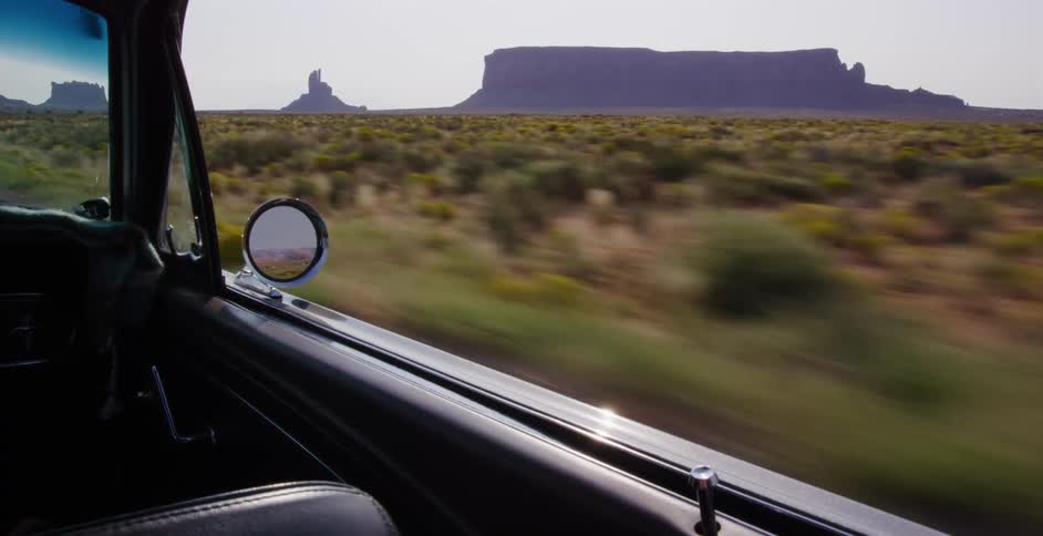 A classic car drives through the iconic Monument Valley desert landscape as seen from the passenger window