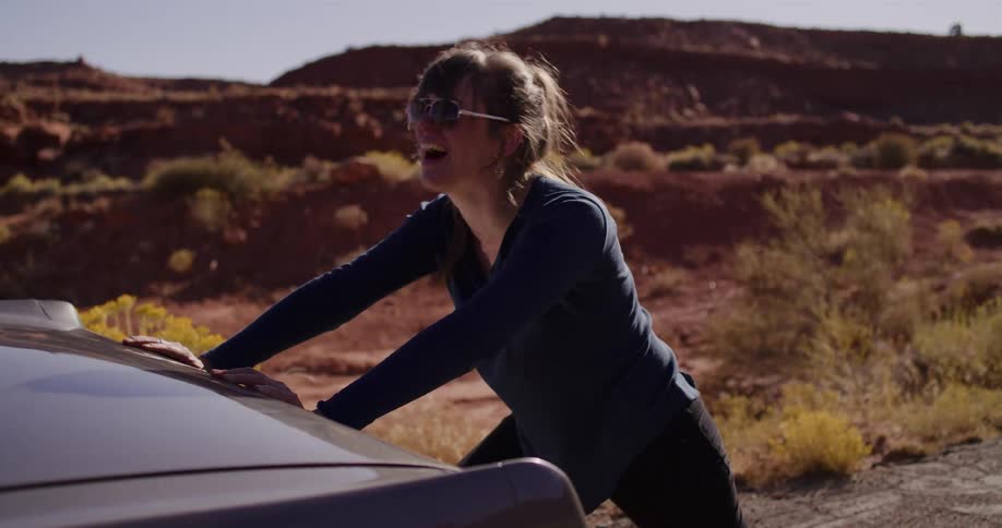A woman struggles and laughs as she tries to push her broken down car on the side of a desert road