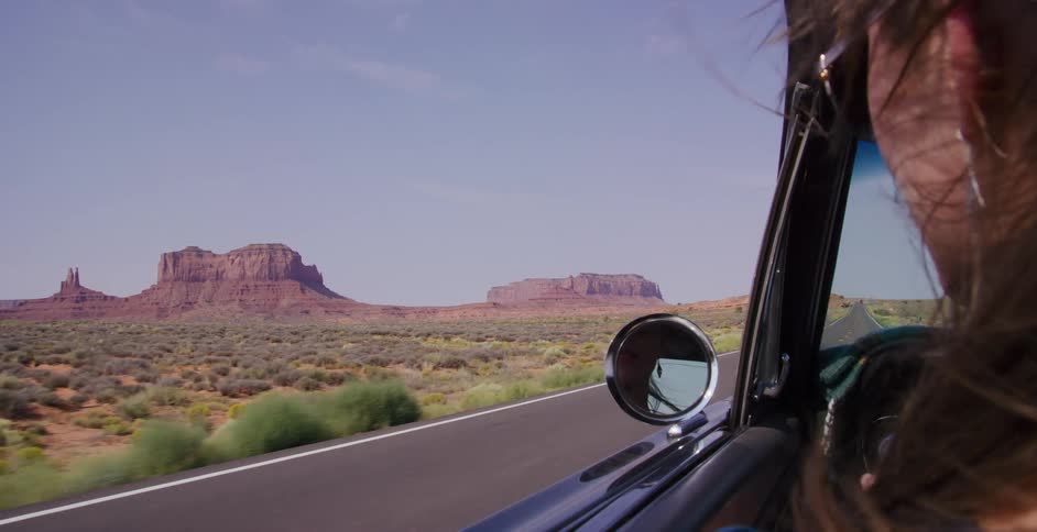 A woman with her hair blowing in the wind enjoys the view of Monument Valley from a classic car on a desert road trip