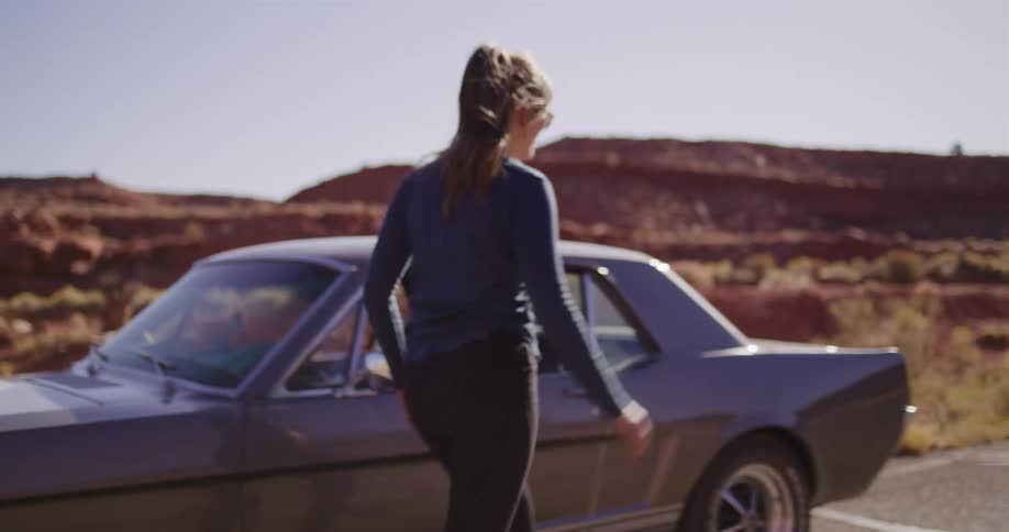 In slow motion, a woman with a ponytail closes the hood of a vintage car and walks to the driver's door in the desert