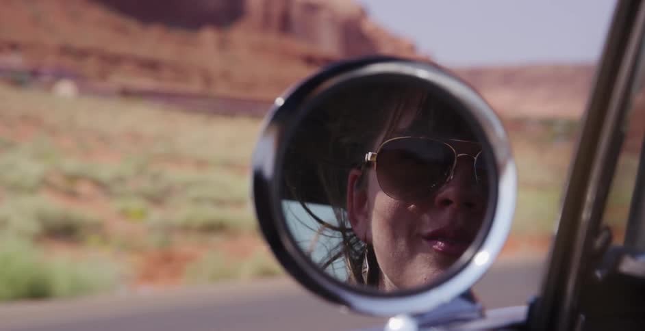 A woman's reflection in a classic car's side-view mirror captures the freedom of a road trip through the desert, with the wind blowing through her hair