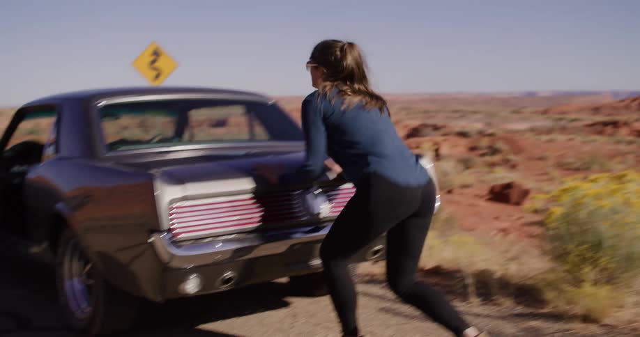 A woman playfully runs and then struggles to push her broken down classic car on a desert road