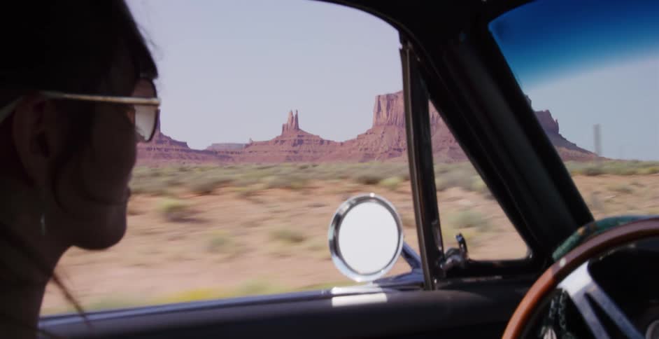 A woman in a classic car drives through the iconic desert landscape of Monument Valley on a sunny day