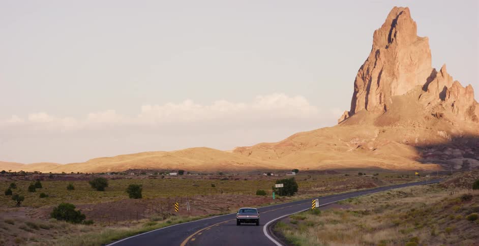 A vintage American muscle car drives along a scenic desert highway towards a massive rock monolith at golden hour