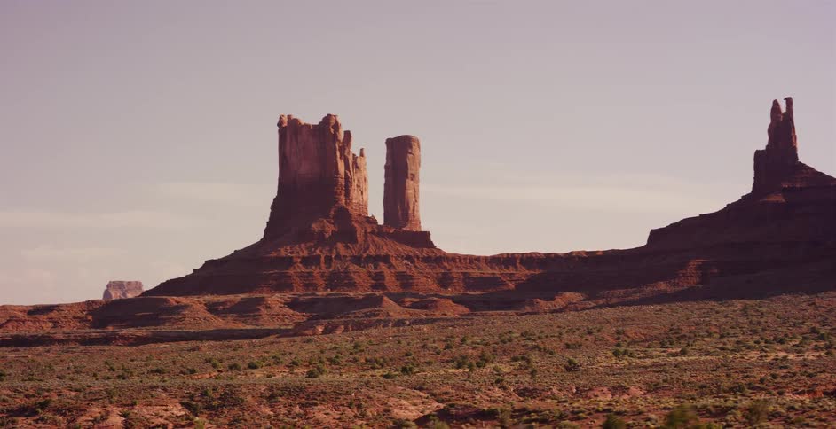 A scenic driving shot reveals the iconic red rock buttes and mesas of Monument Valley under a vast, hazy sky