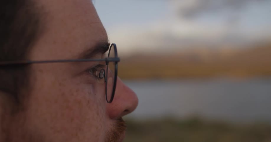 An intimate close-up shot of a thoughtful man wearing glasses as he looks out over a serene landscape at sunset