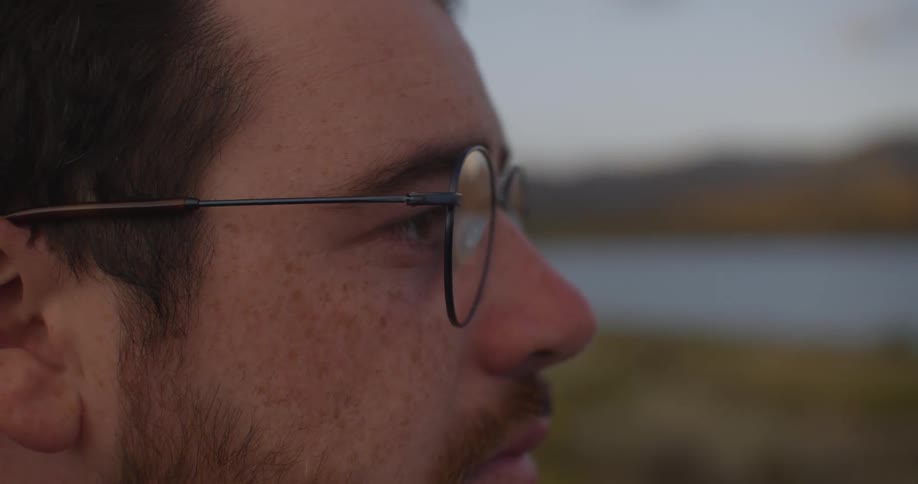 A thoughtful young man with glasses and freckles gazes at a distant lake and mountains at dusk