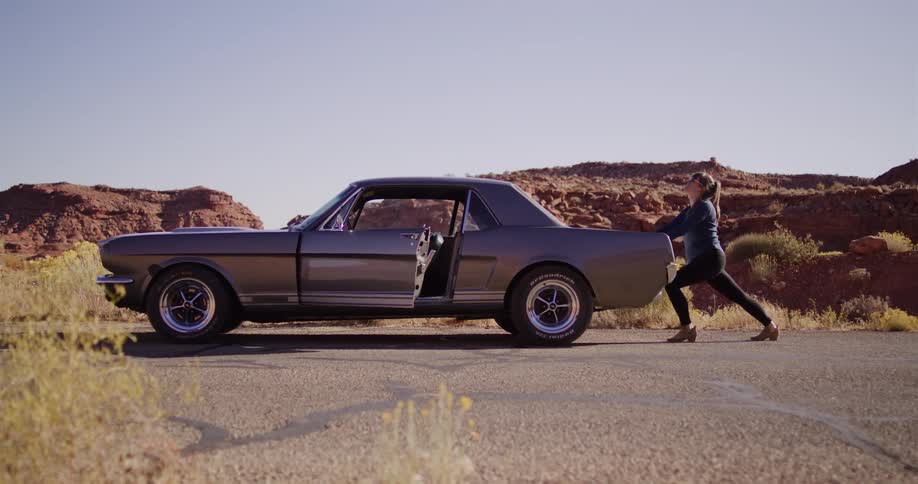 A woman tries to push her broken-down classic muscle car on a desert road and gives up in frustration