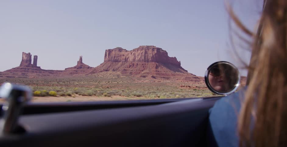The iconic buttes of Monument Valley pass by the window of a classic car, with the female passenger's reflection visible in the side view mirror