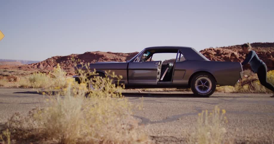A determined woman in slow motion pushes her classic Mustang that has broken down on a desert road, then gets in as it starts to roll downhill