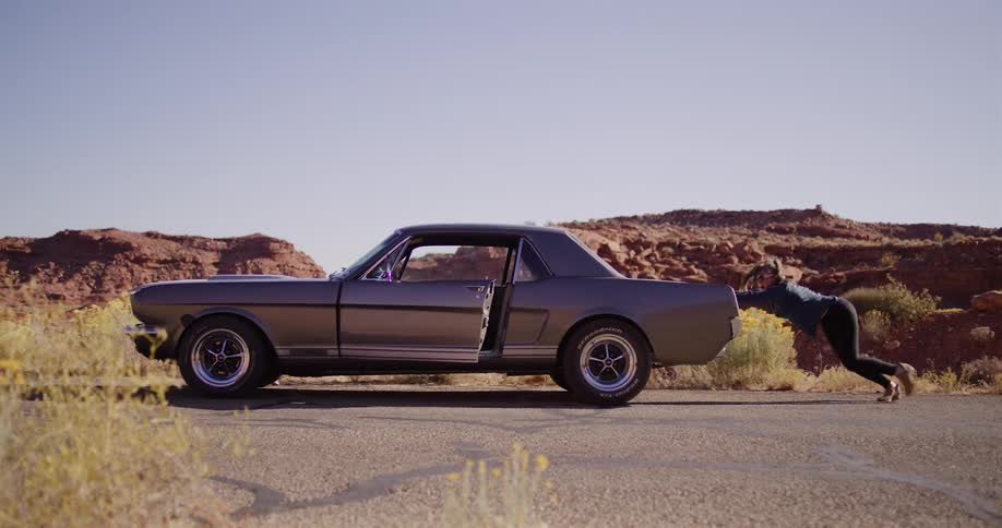 A determined woman single-handedly pushes her broken-down classic Mustang along a deserted road in the arid desert landscape