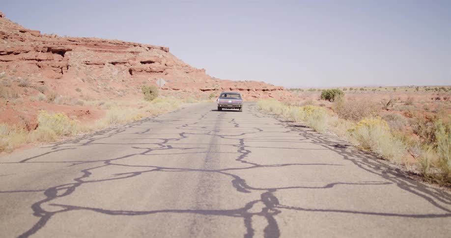 A classic car drives away down a long, cracked desert road surrounded by red rock formations