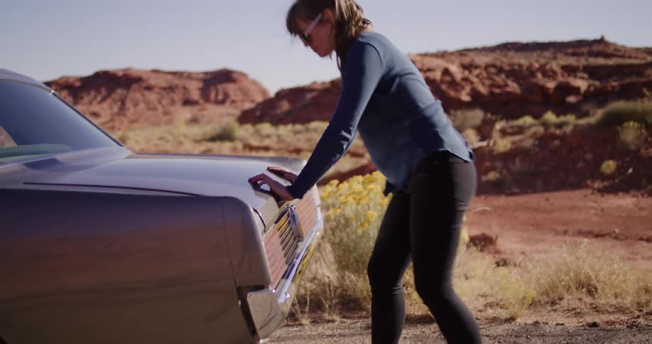 A woman struggles in slow motion to push her broken-down vintage car along a desolate desert road