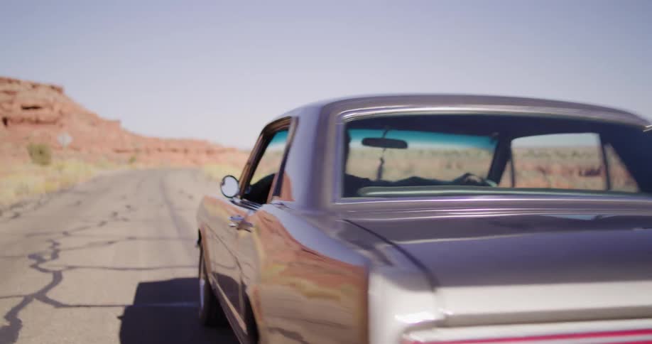 A woman closes the door of a classic grey Mustang and drives away down a cracked road in the desert