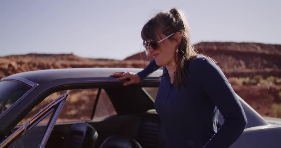 A woman in sunglasses gets out of her classic car on a desert road trip, leaning against it and laughing in slow motion as the sun flares