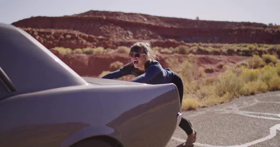 A determined young woman pushes her broken-down classic car along a desert highway with red rock formations in the background
