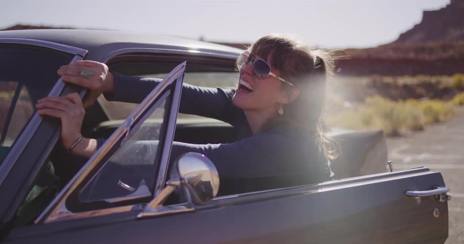 A joyful woman laughs as she gets out of her vintage car, enjoying a sunny road trip through the desert in slow motion