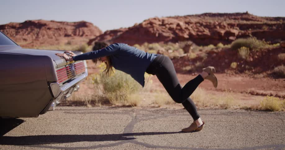 A frustrated woman struggles in slow motion to push her stalled classic car on a deserted desert road before giving up