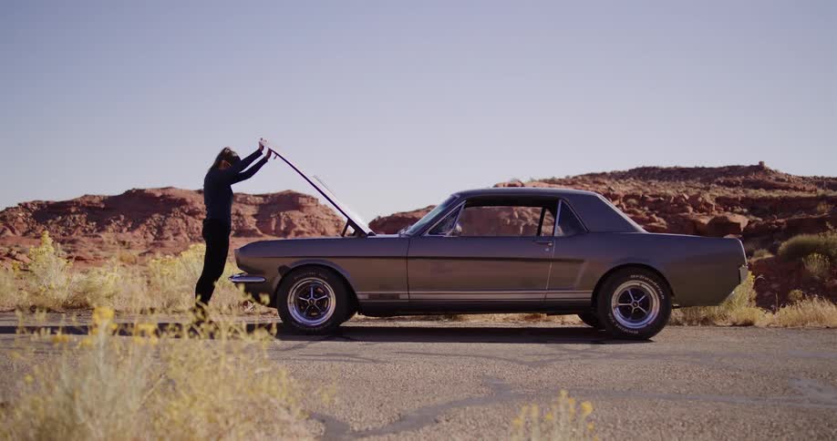 A woman gets out of her classic muscle car after it breaks down on a desert road, opens the hood to investigate, and then gives up in frustration