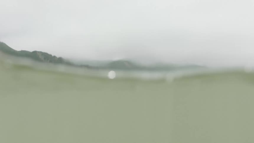 Immersive water-level view of a female surfer paddling out to sea on a misty day with mountains in the background