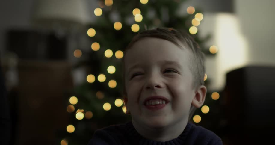 A young boy sitting in front of a christmas tree smiling and laughing