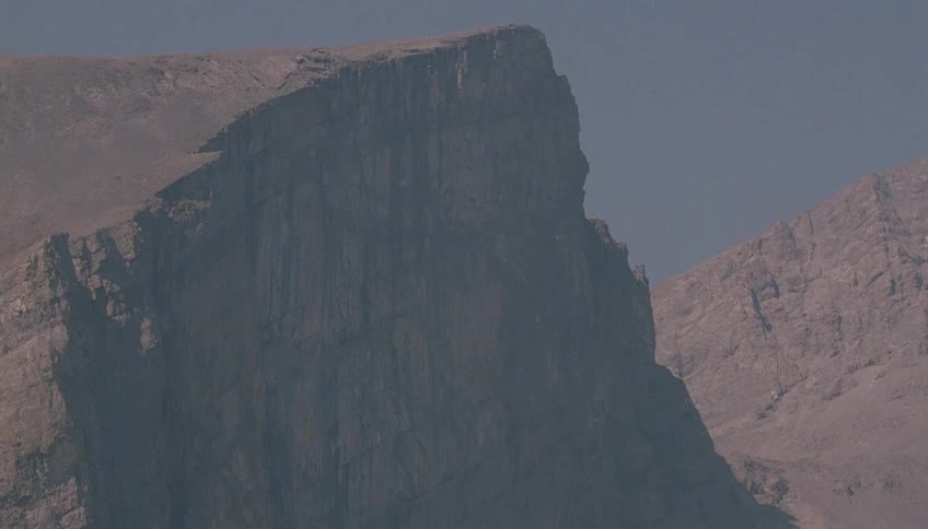 A telephoto shot captures the immense, flat-topped peak of a rocky mountain range under a hazy sky