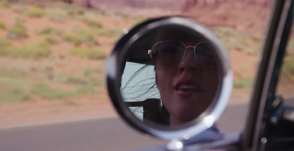 Slow motion reflection of a smiling woman in sunglasses driving through the desert, seen in the car's side mirror