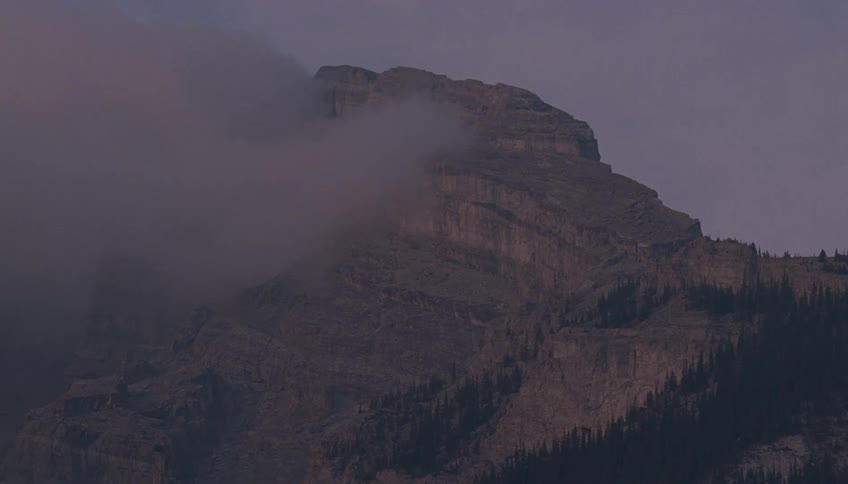 A rugged, rocky mountain peak is enveloped by swirling clouds at dusk