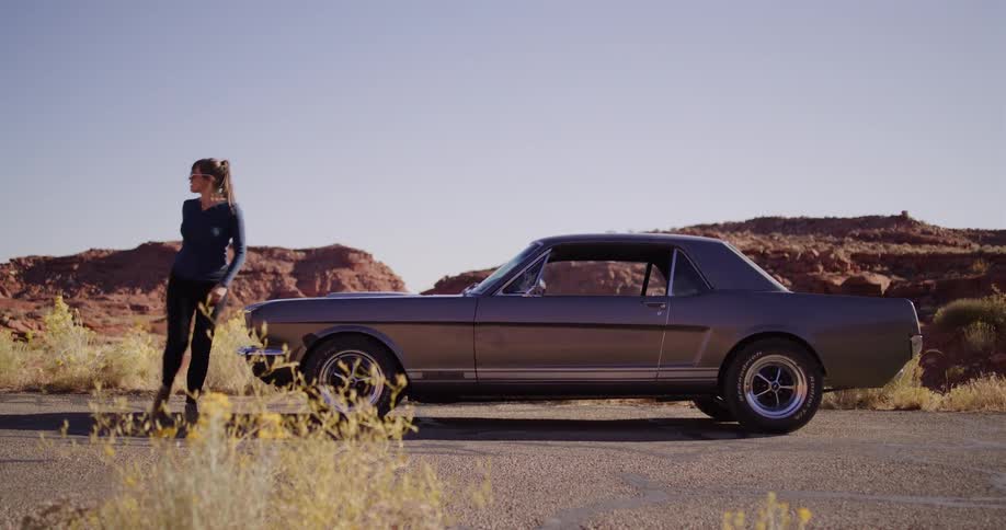 A woman gets out of her classic car, attempts to push it, then opens the hood to investigate why it has broken down on a desert road