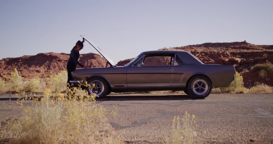 A woman gets out of her broken-down classic car on a desert road and inspects the engine under the hood