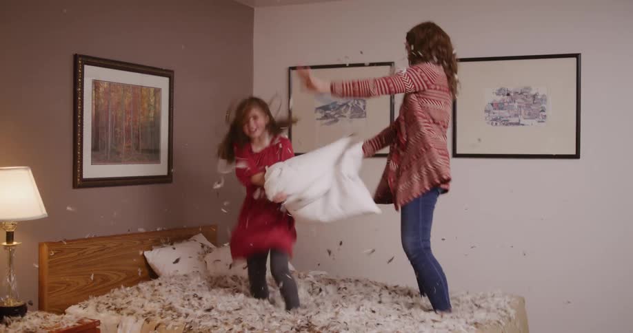 A mother and daughter jump on a bed covered in feathers while having a pillow fight in slow motion