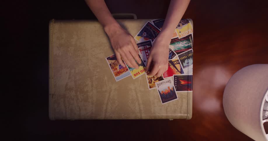 A traveler's hands carefully arrange a collage of souvenir postcards from various destinations onto a vintage suitcase