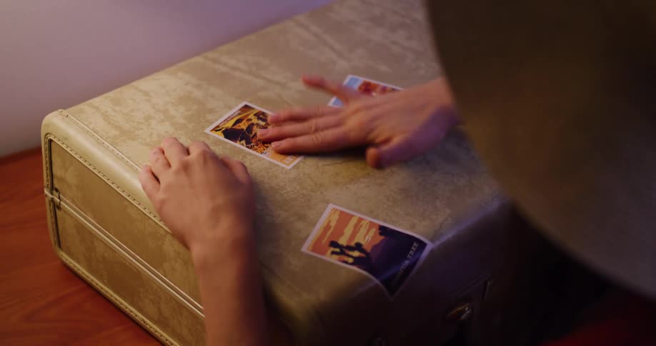 An over-the-shoulder view shows a person's hands carefully applying a travel sticker to a vintage suitcase
