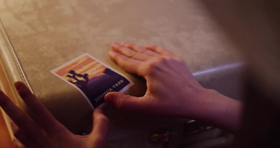 A traveler carefully applies a Joshua Tree souvenir sticker onto a vintage suitcase, commemorating their journey